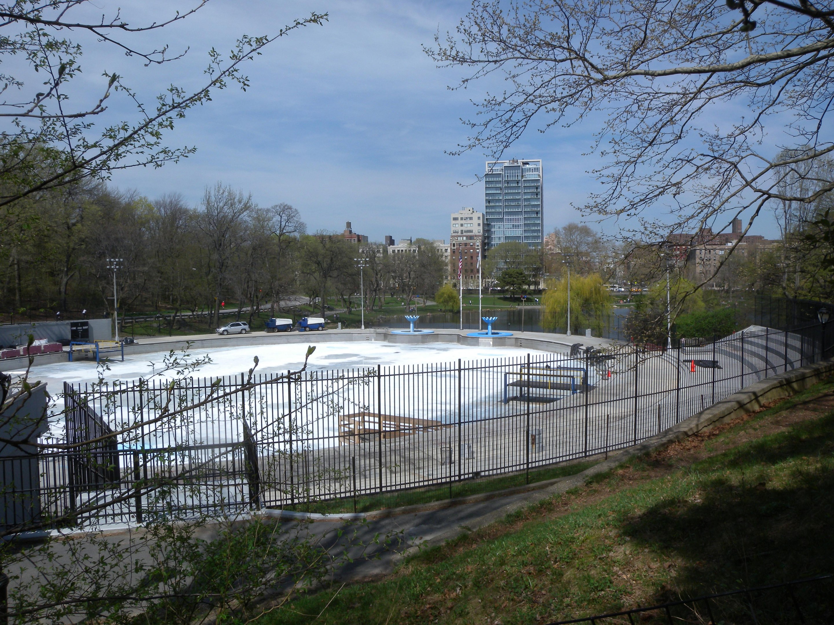 Eisbahn in einem Park umgeben von einem Metallzaun, Gras, Bäumen, Fahrzeugen, Straßenlaternen, einem Gewässer, Gebäuden und einem bewölkten Himmel.
