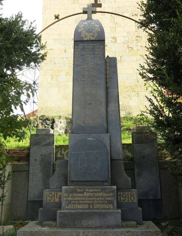 Ein steinernes Denkmal mit einem Kreuz, gewidmet den deutschen Soldaten, die im Zweiten Weltkrieg starben, steht vor einer Kirche, umgeben von Bäumen und einer Mauer.