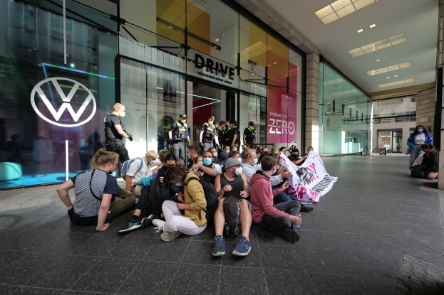 Eine Gruppe von Menschen sitzt vor einer Volkswagen-Vertretung mit Glaswänden auf dem Boden und hält eine Fahne mit Text, unter Deckenleuchten, scheinbar protestierend.