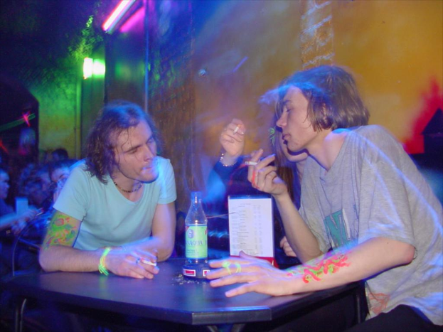 Ein Tisch mit einer Flasche, Speisekarte und Aschenbecher in der Mitte einer Indoor-Szene, mit drei Personen, die Zigaretten rauchen.