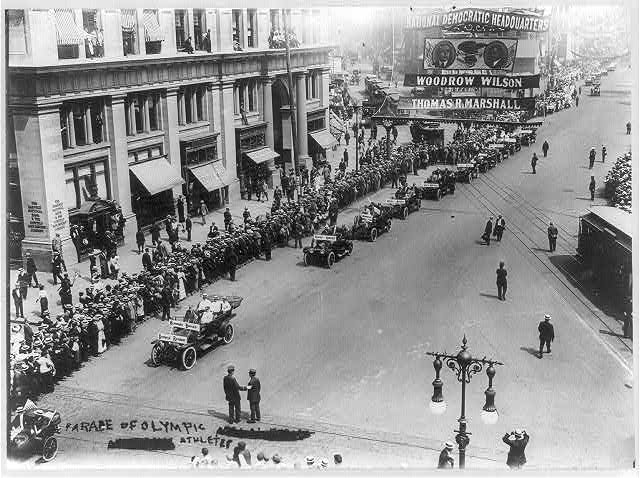 Ein altes Schwarz-Weiß-Foto eines Umzugs auf einer Stadtstraße mit Menschen, Fahrzeugen, Straßenlaternen, einem Schild, Gebäuden und einem bewölkten Himmel.
