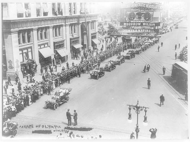 Ein altes Schwarz-Weiß-Foto eines Umzugs auf einer Stadtstraße mit Menschen, Fahrzeugen, Straßenlaternen, einem Schild, Gebäuden und einem bewölkten Himmel.