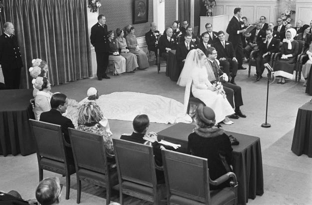 Schwarzes und weißes Foto einer Hochzeitszeremonie mit sitzenden Gästen, einem Mann mit Mikrofon, einer Frau mit einem Strauß, einem Podium mit Mikrofon, Blumengestecken, einem Vorhang, einem gerahmten Foto an der Wand und einer Tür im Hintergrund.