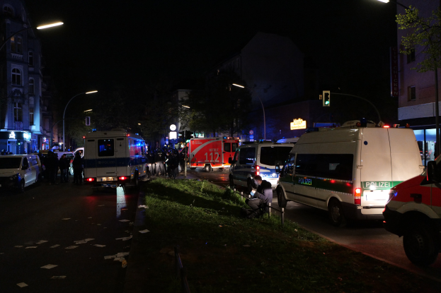 Eine Gruppe von Polizeiwagen steht nachts am Straßenrand, mit Menschen auf dem Gehweg, Gras, Polen, Lichtern, Verkehrsampeln, Schildern, Bäumen, Gebäuden mit Fenstern und dem Himmel im Hintergrund.