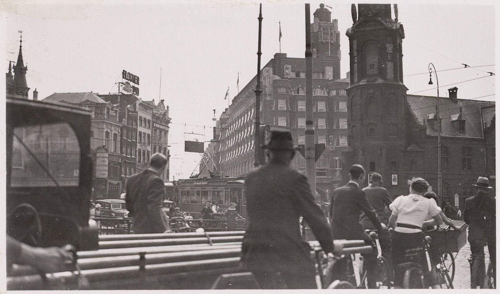 Schwarzes und weißes Foto von Menschen, die auf einer Stadtstraße in Amsterdam im frühen 20. Jahrhundert Fahrräder fahren, mit Gebäuden, Pfählen und Drähten, die die Straße säumen, und dem Himmel im Hintergrund.