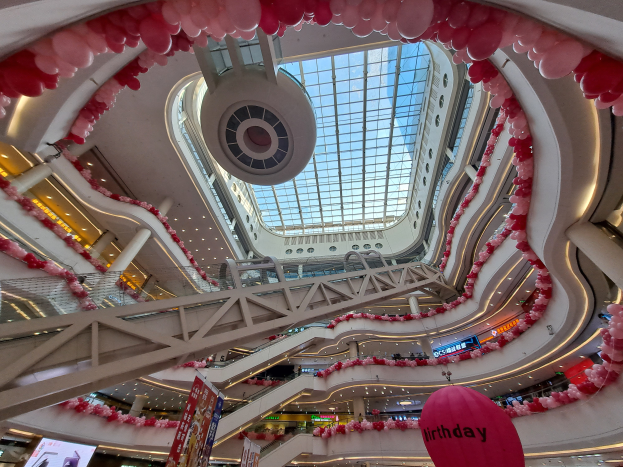 Ein Einkaufszentrum-Innenraum, der für den Valentinstag mit pinken und weißen Luftballons, Rolltreppen, Geländern, Texttafeln, Lichtern und einer Glasdecke dekoriert ist.
