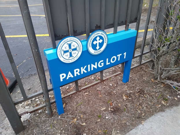 Ein blau-weißes Schild mit der Aufschrift 'Parkplatz 1' steht vor einem Metallzaun, im Hintergrund eine Straße und rechts ein Baum.