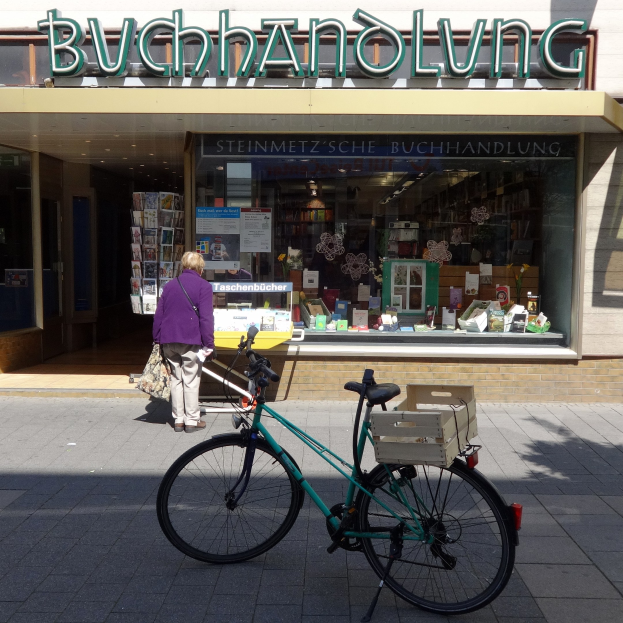 Eine Frau steht neben einem geparkten Fahrrad vor einem Laden, mit einem Fahrradrad im Vordergrund und Büchern und anderen Gegenständen durch die Glasscheibe des Ladens sichtbar.