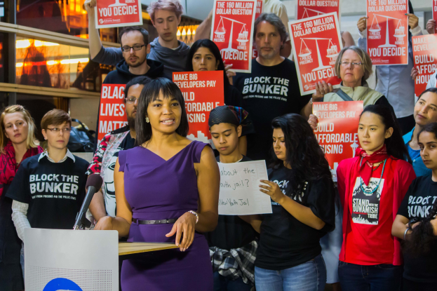 Eine Black-Lives-Matter-Aktivistin spricht in ein Mikrofon an einem Rednerpult zu einer Menge, die Schilder hält, mit Bühnenlicht und Equipment im Hintergrund.