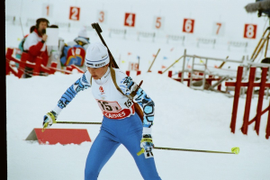 Eine Frau in einer blauen und weißen Kleidung, die eine schneebedeckte Anhöhe mit Ski und Skistöcken hinunterfährt, mit Menschen, einer Kamera auf einem Stativ und einer Nummerntafel im Hintergrund.