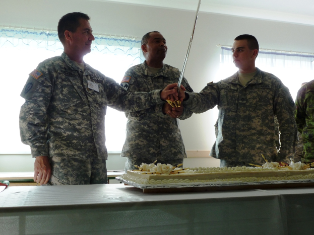 Ein großer Kuchen auf einem Tisch mit mehreren Soldaten davor, drei halten ein großes Messer, und eine Wand mit zwei Fenstern im Hintergrund.
