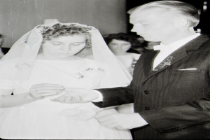 Ein Schwarz-Weiß-Foto einer Braut in einem weißen Kleid und Schleier und eines Bräutigams in einem Anzug beim Austausch der Eheringe, mit ein paar Menschen im Hintergrund.