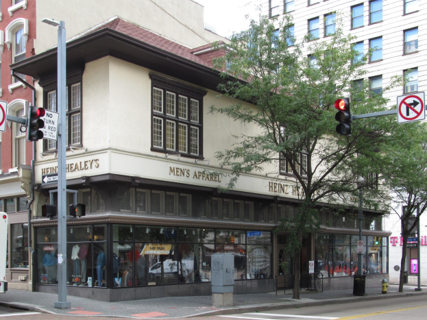 Eine belebte Straßenkreuzung mit einem geparkten Bus, Gebäuden, Bäumen, Laternen, Ampeln, Schildern und einem Kaufhaus mit Schaufensterpuppen in Kleidern vor einem sichtbaren Himmel.