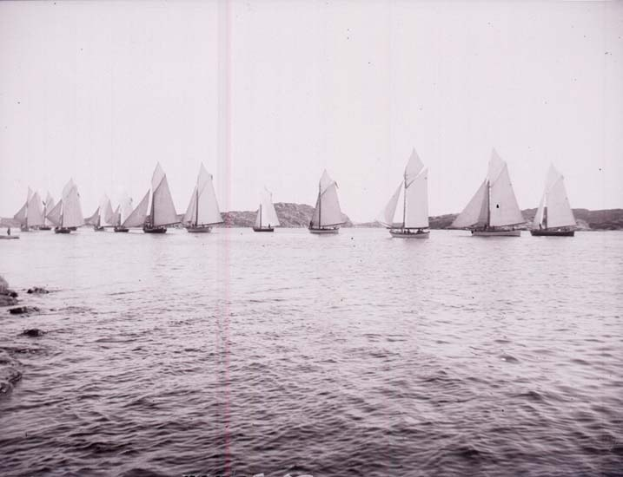 Schwarzes und weißes Foto von Segelbooten beim Rennen auf dem Wasser mit Hügeln im Hintergrund und einem klaren Himmel, beschriftet als New York Yacht Club.