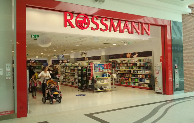 Ein Rossmann-Laden in einem Einkaufszentrum mit Menschen, einer Person mit einem Kinderwagen, und Regalen mit Produkten unter Deckenbeleuchtung und Beschilderung.