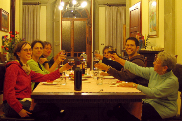Eine Gruppe von Menschen sitzt an einem Tisch und hält Weinpokale in den Händen, mit Flaschen, Tellern, einem Foto auf der Wand und Vorhängen im Hintergrund.