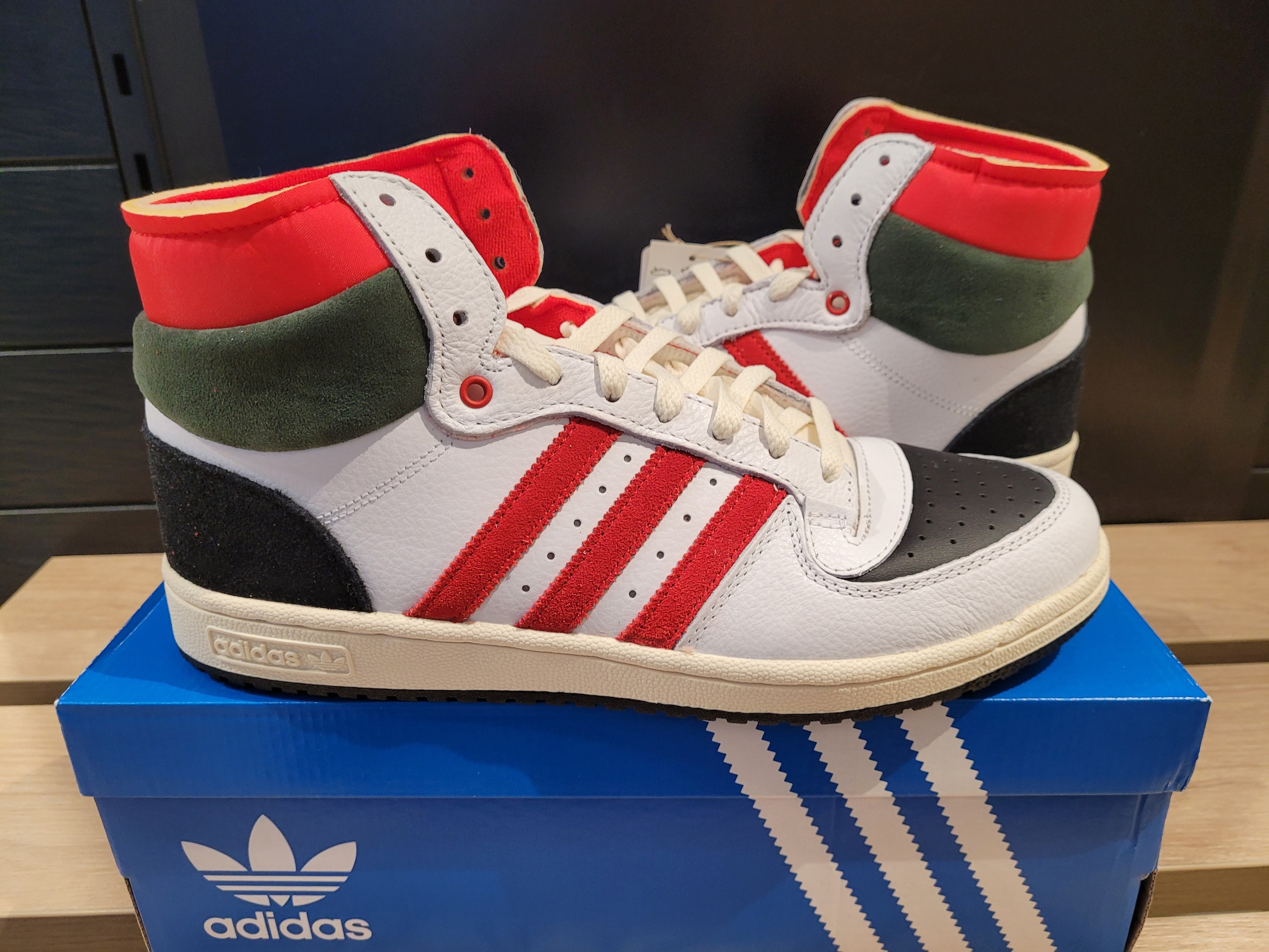 Weiße, rote, grüne und schwarze Adidas Originals Top Ten Hi-Schuhe auf einer blauen und braunen Kartonschachtel auf einem cremefarbenen Tisch vor einer schwarzen Wand.