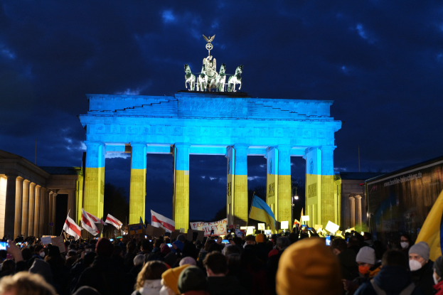 Eine Menge steht vor dem Brandenburger Tor in Berlin, hält Fahnen und Schilder, mit einem Banner auf der rechten Seite, das protestbezogene Texte anzeigt.