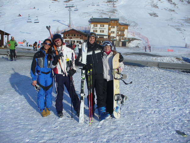 Vier Personen posen für ein Foto mit Skater-Pads und Werkzeugen, mit Gebäuden und anderen Menschen im Hintergrund, vor einer schneebedeckten und vereisten Berglandschaft.