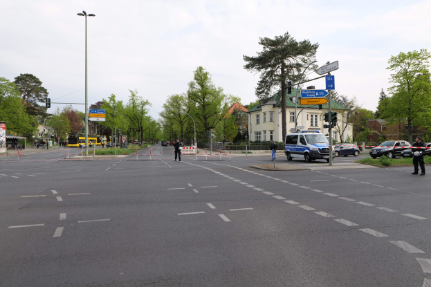 Eine Kreisverkehres mit einem parkenden Polizeiauto, Fahrzeugen auf der Straße, Fußgängern auf dem Gehweg, Verkehrsampeln, Bäumen, Gebäuden mit Fenstern und einem bewölkten Himmel.