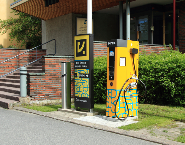Eine Elektrofahrzeug-Ladestation an der Straße, umgeben von Grün und einem Gebäude mit Fenstern und Säulen, mit Stufen und Geländern, die hinaufführen.