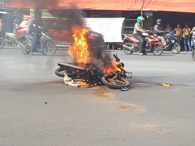 Motorrad in Flammen an der Straße mit einem Auto und einem Van im Hintergrund, umgeben von Menschen, einer Fahne und einem Zelt.