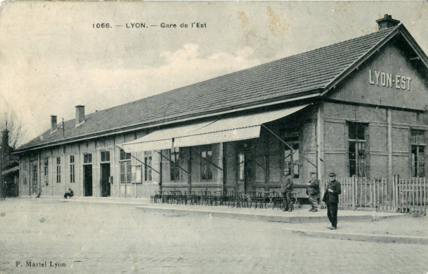 Ein altes Schwarz-Weiß-Foto eines Bahnhofs in Lyon, Frankreich, mit einem Gebäude mit Fenstern, einem Dach, einem Zaun, einer Gruppe von Menschen auf dem Boden, einigen Stühlen, einer Person auf einer Bank sitzend, einigen Bäumen und einem bewölkten Himmel.