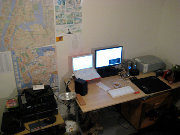Ein Raum mit einem Tisch, auf dem ein Laptop, Lautsprecher, Monitor, Tastatur, Ball, Maus, Scanner und Papiere liegen, und einem weiteren Tisch mit Elektronikgeräten, sowie Charts und Postern an der Wand dahinter.