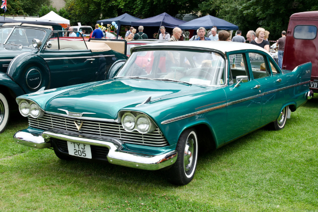 Vintage-Autos auf Gras geparkt mit Menschen dahinter, Zelte im Hintergrund, Bäume oben und eine Flagge oben links.