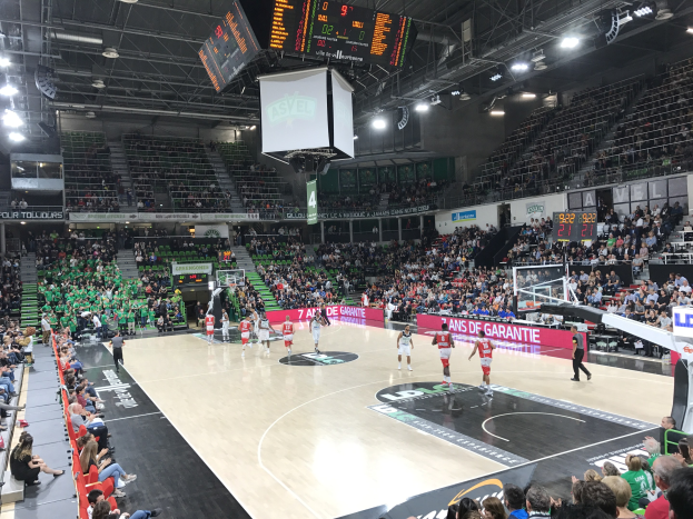 Basketballspiel in einer großen Arena mit Zuschauern, Anzeigetafeln mit "Asvel vs Galatasaray" und Decke mit Beleuchtung und Bildschirm.