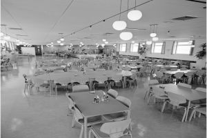 Ein geräumiger Speiseraum mit Reihen von Tischen und Stühlen, verstreute Flaschen und Gegenstände, Deckenleuchten, Fenster auf der rechten Seite und Topfpflanzen überall.