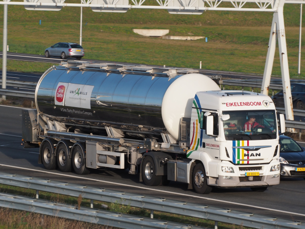 Ein großer Tanklastwagen und ein Auto fahren nebeneinander auf einer Autobahn, mit sichtbaren Insassen im Lastwagen und Gras, Strommasten und anderen Gegenständen im Hintergrund.