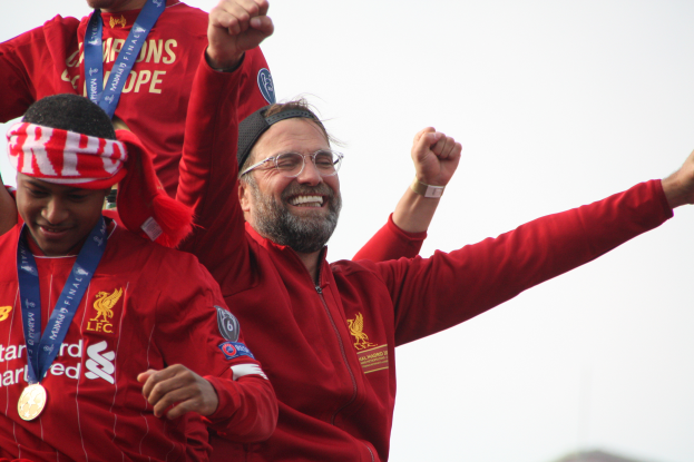 Liverpool-Trainer Jürgen Klopp feiert in rotem Jackett mit seinen Spielern den Sieg der UEFA Champions League, umgeben von zwei medaillentragenden Personen und dem Himmel im Hintergrund.