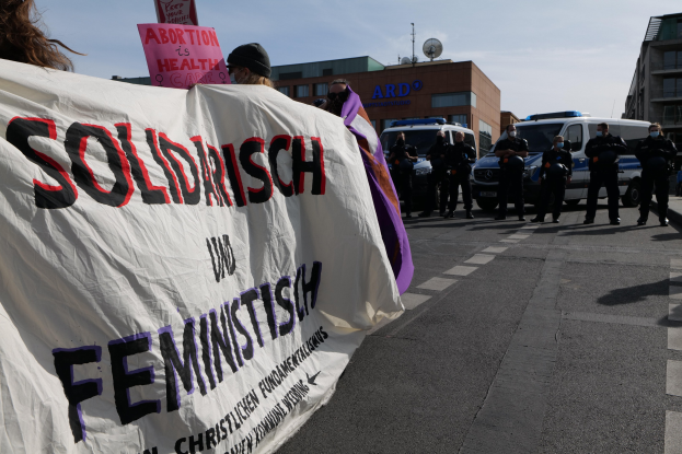 Eine Gruppe von Menschen marschiert auf einer Straße, hält ein Banner mit der Aufschrift "Solidarität und Feminismus" und hat geparkte Fahrzeuge und Gebäude im Hintergrund bei klarem blauem Himmel.