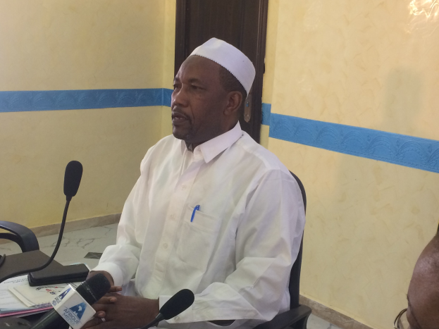 Ein Mann in weißem Anzug und Hut sitzt an einem Tisch mit Mikrofonen, spricht, während Papiere und Gegenstände auf dem Tisch liegen, mit einer Person mit Brille rechts von ihm in einem Raum mit Wand und Tür.