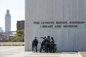 Eine Gruppe von Polizisten in blauen Uniformen und Helmen steht vor der Lyndon Baines Johnson Bibliothek und dem Museum, mit Fahrrädern in den Händen, vor einer textbeflaggten Wand, Treppen, Bäumen, Gebäuden und einem klaren blauen Himmel im Hintergrund.