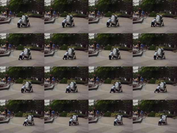 Ein Kind geht auf einer Straße und zieht einen Wagen, mit zwei Personen, die links Fahrräder halten, Bäumen im Hintergrund und einem Gebäude auf der linken Seite.