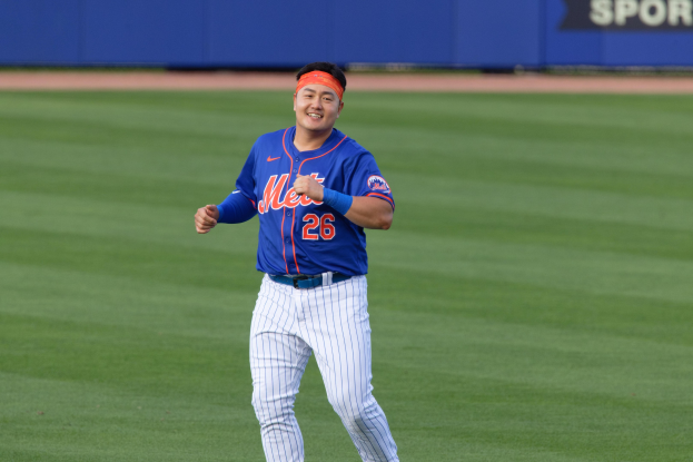 Ein grinsender Baseballspieler in einem blauen T-Shirt, weißen Hosen und einem orangen Stirnband rennt über ein Feld mit einer blauen Tafel im Hintergrund.