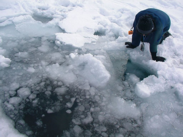 Eine Person kniet rechts im Bild auf dem Schnee und schaut auf das Eis in der Mitte eines Gewässers.