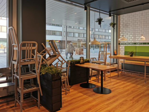Ein Berliner Restaurant mit Holztischen und -stühlen vor einem großen Fenster, das städtische Elemente wie Passanten, Gebäude und Straßenmöbel zeigt.