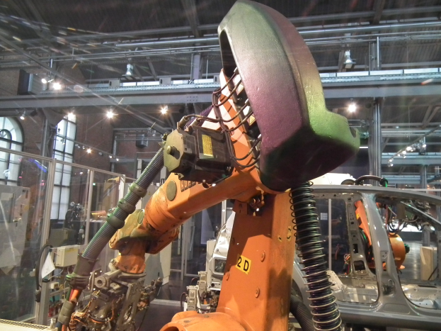 Ein Roboterarm arbeitet an einem Auto in einer industriellen Umgebung mit Metallstangen, Beleuchtung und anderen Maschinen, getrennt durch eine Glaswand von weiterer Hintergrundausrüstung.