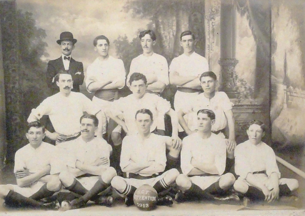 Ein Schwarz-Wei"s-Foto einer Fu"ballmannschaft, mit einigen Spielern, die sitzen und einigen, die stehen, einem Ball in der Mitte und B"umen und einem klaren Himmel im Hintergrund, mit Text unten.