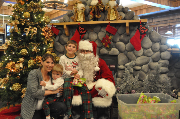 Eine Frau, Kinder und ein als Santa Claus verkleideter Mann mit Brille und Handschuhen stehen neben einem geschmückten Weihnachtsbaum, einer Wand mit Felsen, Spielzeugen, Dekorationen, einem Korb mit Gegenständen und Hintergrundbeleuchtung, während ein Junge ein Päckchen hält.