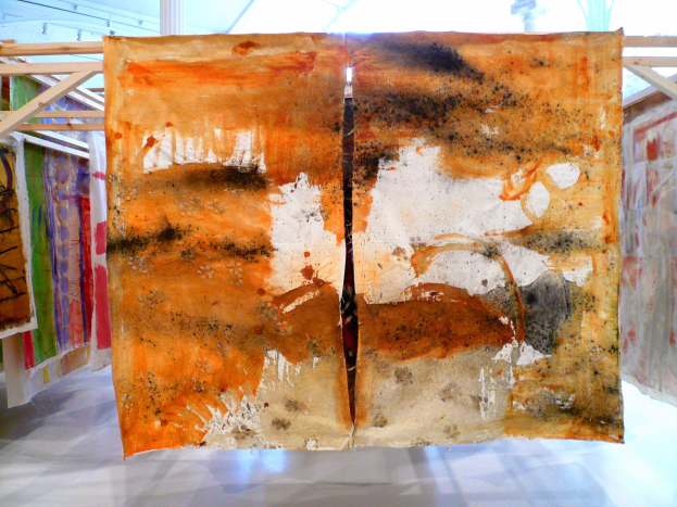 Ein großes, buntes Kunstwerk mit verschiedenen Texturen hängt an einer Wand in einem Raum, mit einer weißen Oberfläche unten und einer Decke oben.