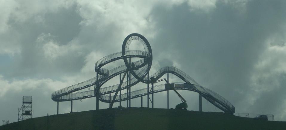 Eine Achterbahn auf dem Höhepunkt eines Hügels mit einem bewölkten Himmel, umgeben von Stützpfählen und Fahrgeschäftsinfrastruktur.
