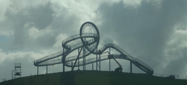Eine Achterbahn auf dem Höhepunkt eines Hügels mit einem bewölkten Himmel, umgeben von Stützpfählen und Fahrgeschäftsinfrastruktur.
