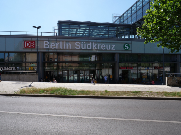 Großes modernes Berliner Südkreuz-Bahnhof mit Glaswänden, umgeben von Straßeninfrastruktur, Fahrzeugen, Fußgängern, Fahrrädern, Bäumen und einem klaren Himmel.