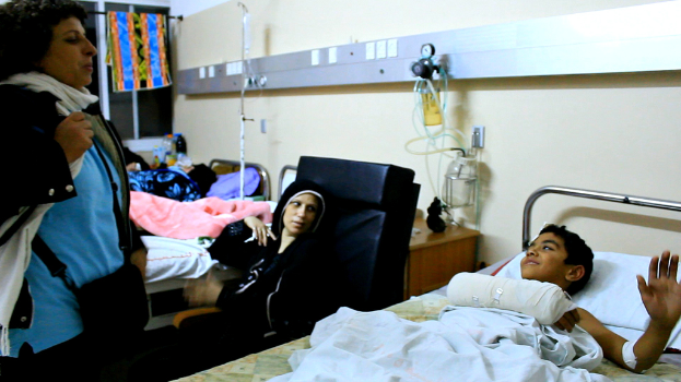 Ein Krankenhauszimmer mit mehreren Betten, an denen Patienten liegen, Infusionsflaschen auf Ständern zwischen den Betten und eine Frau mit einer Tasche in der Nähe eines Patienten.