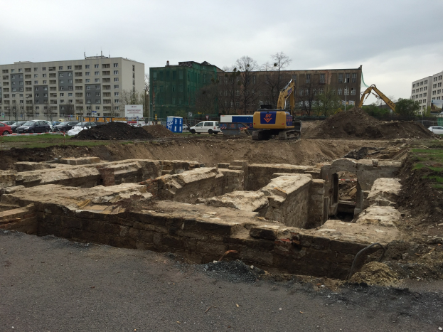 Großer Schutthaufen auf einer Baustelle umgeben von Baggern, Fahrzeugen, Gras, Bäumen, Gebäuden und einer Straße.