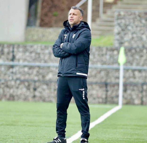Ein Mann steht auf einem Fussballfeld mit verschränkten Armen, trägt eine schwarze Jacke und Hose, mit einem Zaun, Treppe und einer Wand im Hintergrund.